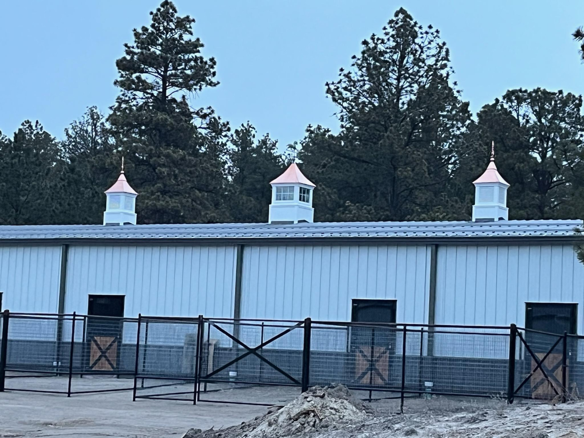 Steel horse barn with white wall panels, copper-topped cupolas and stall runs opening to outdoor paddocks.